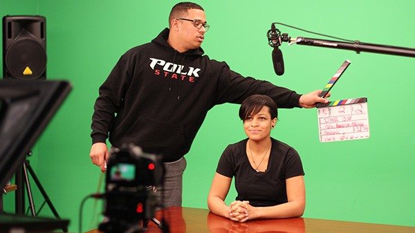 Keith Greene holds out a clapper behind Ashley Bramwell in the green screen studio.